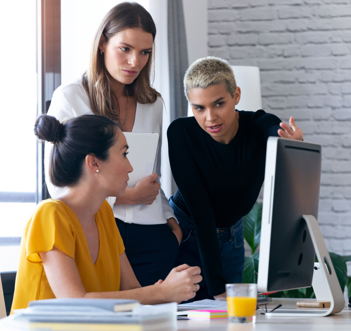 Three people in a discussion around a computer in an office setting, focused on pilot mental health. One person in a yellow top is seated, while two others stand, one holding a notepad and the other gesturing thoughtfully towards the screen.