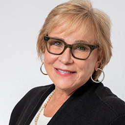 Woman with short blonde hair wearing glasses, hoop earrings, a black jacket, and a white top, smiles against a plain background.